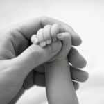 A black and white close-up of a newborn baby's tiny hand and arm resting against a mother's hand